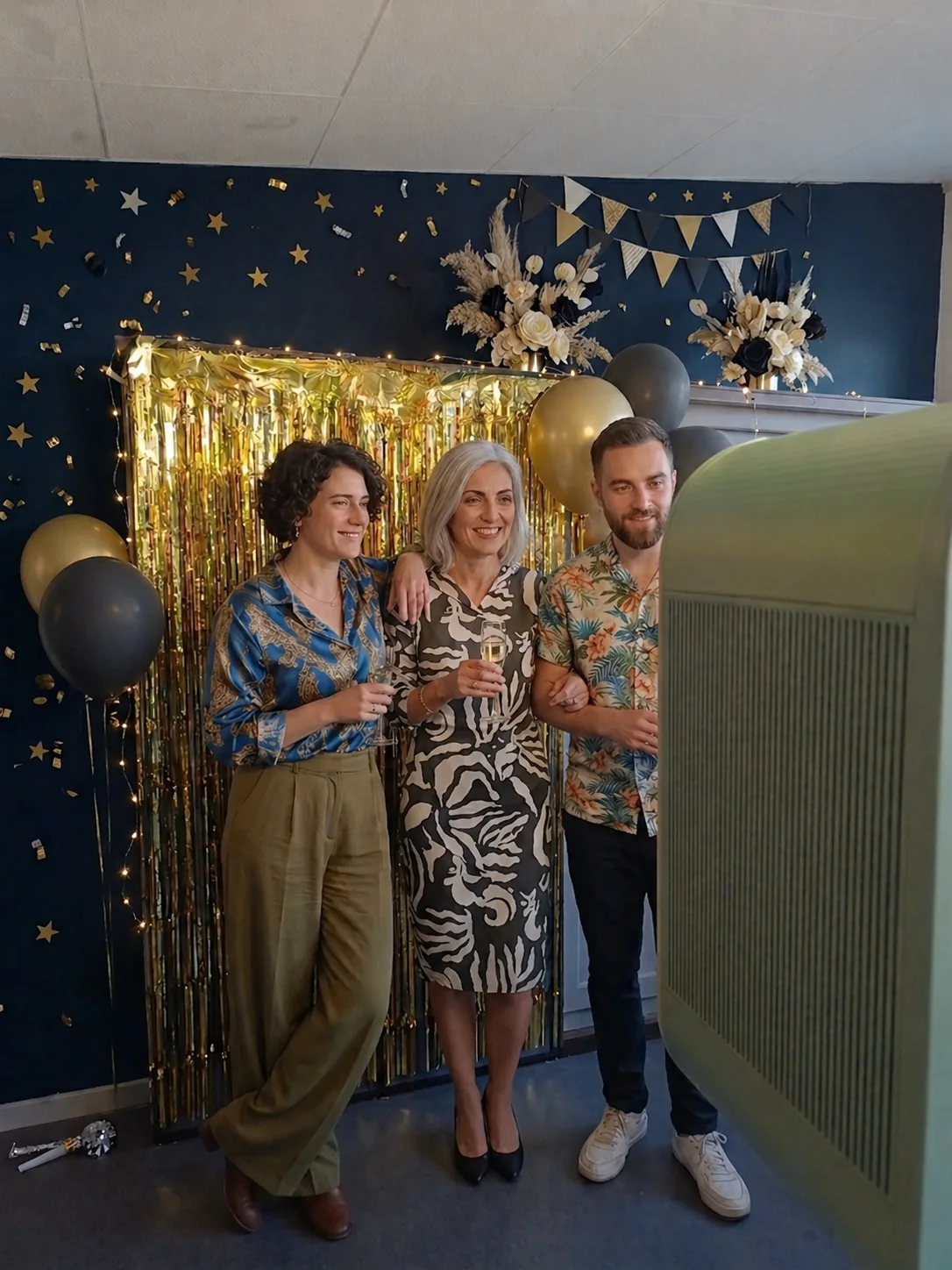 Trois invités souriants, coupes de champagne à la main, posent devant un rideau doré scintillant et la borne photo Burgundy Booth
